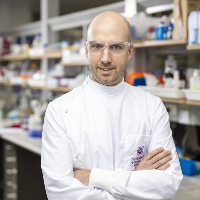 Head shot of Dr Dan Watterson with his arms folded across his chest, and the lab behind 